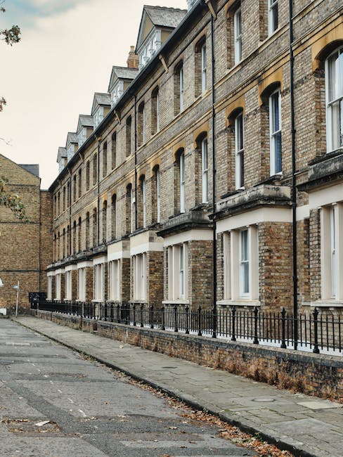 A street view showing a row of Victorian-style terraced houses with brick facades, some painted in light cream and yellow, with bay windows and black wrought iron railings. The street is lined with pa